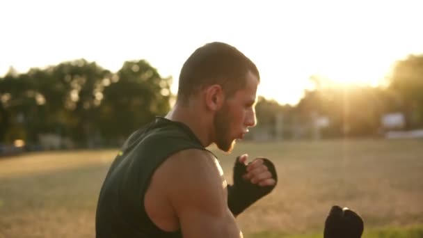 Um jovem boxeador de ataduras pretas trabalhando ao ar livre no estádio - soco. Treino de kickboxing. Sol brilhando no fundo — Vídeo de Stock