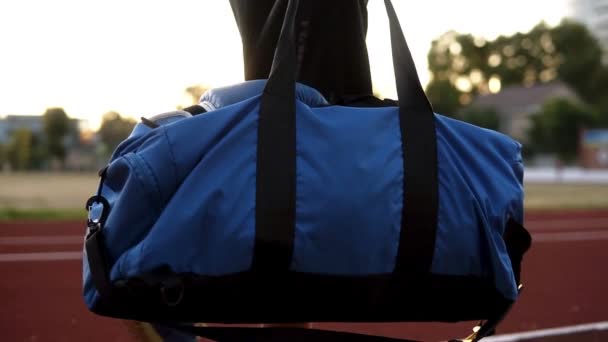 Joven hombre musculoso en camisas negras caminando con bolsa deportiva en la mano por pista de atletismo del estadio. Listo para entrenar, abrir la bolsa y sacar los guantes de boxeo — Vídeo de stock