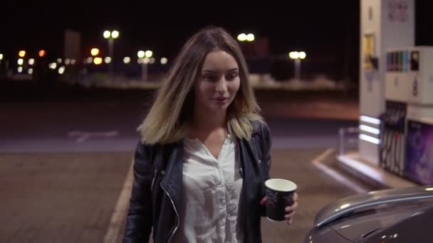 Una joven atractiva abre la puerta del coche mientras sostiene una taza de café y se sienta en un asiento de pasajeros. Gasolinera. De cerca. — Vídeos de Stock