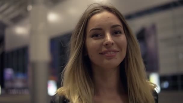 Attraktive junge blonde Frau lächelt und posiert für die Kamera. Verschwommenes Flughafengebäude im Hintergrund — Stockvideo