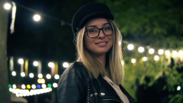 Porträtt av en vacker flicka i snygg hatt, mode glasögon och en svart skinnjacka promenader i parken natt. Ljus av en café lampor på bakgrunden. Blond flicka leende, poserar för kameran — Stockvideo