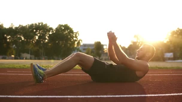 Handsome young man in a black T-shirt pumps the press on the stadium outdoors. Young boxer doing abs and punching exercising on the floor.Sun shines on the background — Stock Video