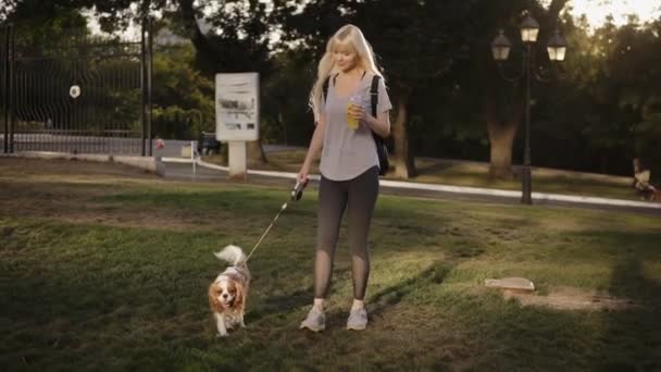 Önden Görünümü ile onun köpek parkta yürüyüş uzun sarı saçlı güzel güleryüzlü kadın. O rahat kıyafetler giymiş ve elinde içki holding — Stok video