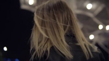 Eğleniyor sarışın genç kadın kot pantolon ve siyah deri ceket geceleri sarı sokak tamponlar üzerinde atlama. Street, beyaz şehir ışık, yaya geçidi arka planda. Görünümünde nadir kamera için poz