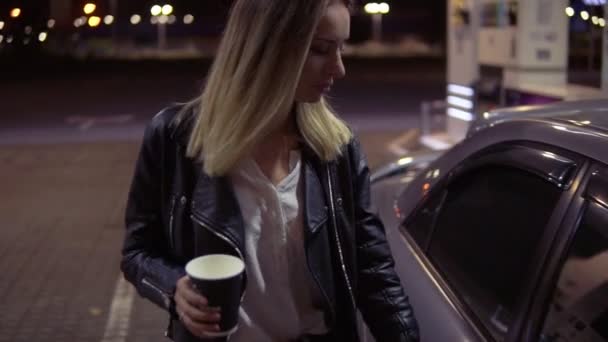Ξανθιά νεαρή γυναίκα ανοίγει την πόρτα του αυτοκινήτου, κρατώντας ένα φλιτζάνι καφέ και κάθεται στο κάθισμα του συνοδηγού. Πρατήριο βενζίνης. Διανυκτέρευση — Αρχείο Βίντεο