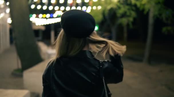 Uma menina bonita em chapéu elegante e uma jaqueta de couro preto caminha pelo parque noturno à luz de um café lâmpadas. Sorrindo, posando para câmera. Vista rara — Vídeo de Stock