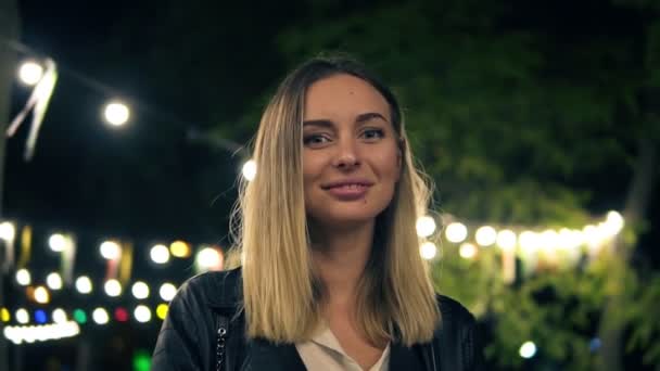 Ritratto di una bella ragazza in giacca di pelle nera e camicetta bianca in piedi nel parco notturno. Luce di un caffè lampade colorate sullo sfondo. Ragazza bionda sorridente, guardando la fotocamera — Video Stock