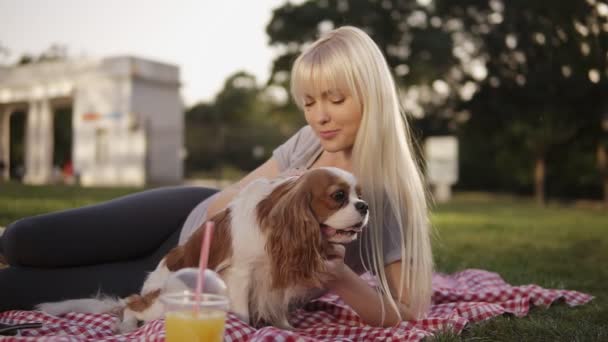 Vue rapprochée d'une femme blonde aux cheveux longs allongée sur un sol dans le parc sur une litière à carreaux et caressant son petit chien. Fond flou, tasse en plastique avec boisson au premier plan — Video