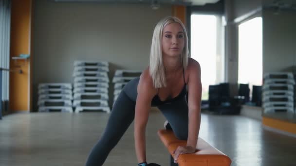 Повільні кадри привабливої молодої жінки, що тренується з гантелями, що спираються на коліно на лавку в порожньому тренажерному залі з дзеркалами. Вид спереду — стокове відео