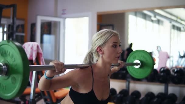 Vista lateral de una chica rubia practicando sentadas usando una barra con pesas verdes. Fondo borroso de gimnasio con equipo y espejo — Vídeos de Stock