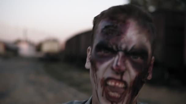 Portrait of a male zombie with bloody teeth and wounded face screaming and shouting. Halloween, filming, staging concept. Blurred railway wagons on the background — Stock Video