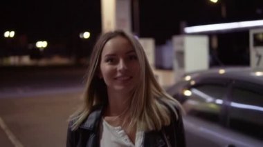 Gülümseyen kadın caual benzin istasyonunda, araba arka plan giriyor ve poz romantik. Önden görünüm. Gece vakti