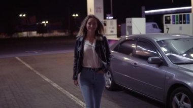 Gülümseyen kadın caual benzin istasyonunda, araba arka plan giriyor ve poz romantik, portresi. Önden görünüm. Gece vakti