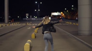 Mutlu sarışın kadın geceleyin açık havada eğleniyor. Havaalanı binasının yanında. Çalışan, immitating uçak, tamponlar üzerinde atlama. Kot pantolon ve siyah ceket giyiyor. Görünümünde nadir