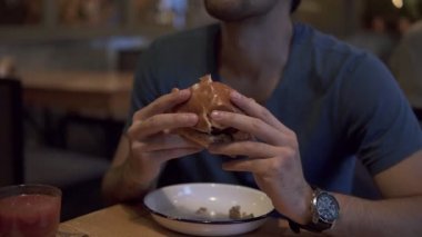 Mavi tişörtlü bir kafede hamburger yemek yerken esmer bir adam. Lezzetli hamburger ile iki el ele tutuşarak, yeme. Bulanık kafe iç arka planda