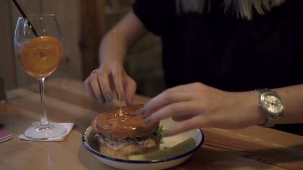Bir hamburger yeme genç bir kadının görüntüleri kapatın. Eller, sonra onu tutan burger iterek genç kadın portresi. Kokteyl masanın üzerine bir bardak. Sağlıksız beslenme, hızlı yağ gıda — Stok video