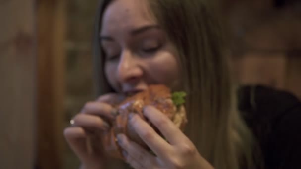 Kopfschuss einer hungrigen Frau, die in einem Café einen Hamburger isst. Porträt einer jungen Frau, die Burger isst und sich in den Händen hält. Ungesunde Ernährung, Fast-Food-Konzept — Stockvideo