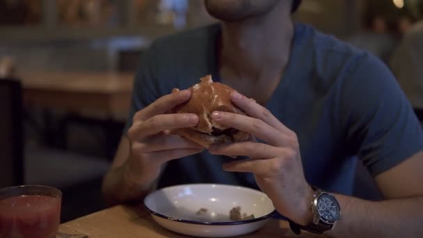 En brunett mannen i blå T-tröja med burger middag i ett café. Äta en välsmakande hamburgare, hålla med två händer. Suddig café inredning på bakgrunden — Stockvideo