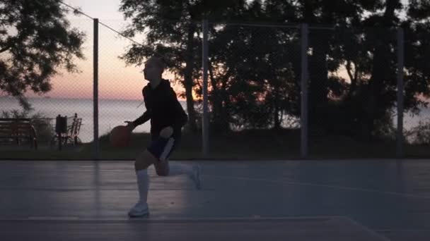 Jonge vrouw basketbal speler opleiding buiten op een basketbalveld. Ochtend schemering — Stockvideo