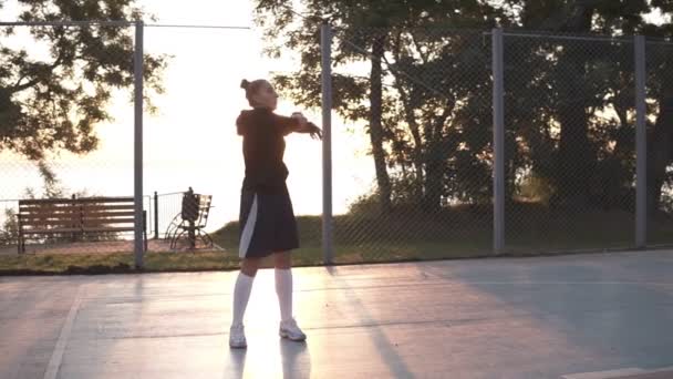 Kaukasische sportieve jongedame in hoodie en shorts aan het warmrijden in de ochtend op het basketbalveld. Zon schijnt op de achtergrond — Stockvideo