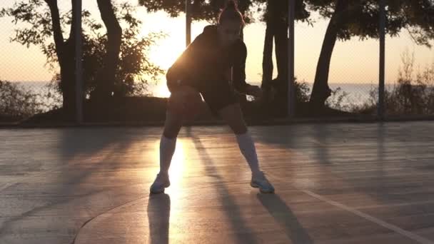 Молодая кавказская баскетболистка дриблинг и практикует навыки обращения с мячом на корте. Утренний закат, солнце светит на заднем плане — стоковое видео