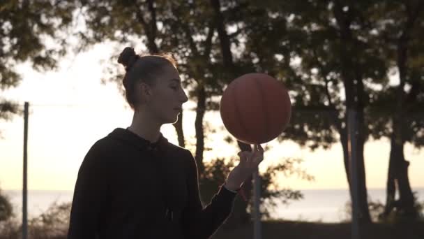 Menina bonita com capuz preto girando uma bola de basquete no dedo ao ar livre. Árvores e sol brilha no fundo — Vídeo de Stock