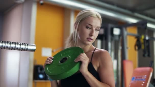 Deportiva chica sacar los pesos de una barra después de entrenar en el gimnasio. Colocar ruedas de barra verde en un soporte — Vídeos de Stock