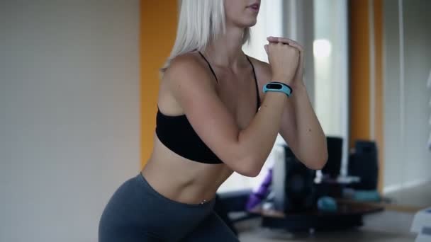 Молодая привлекательная блондинка сидит в спортзале. Женщины профессионально практикуют приседания, тренируются в спортзале. Ношение серых леггинсов и черный лифчик — стоковое видео