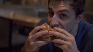 Restoranda lezzetli hamburger yeme mutlu gülümseyen genç adam. Fast food Cafe, Burger yığılmış aç genç adam bir şeyler yapar ve çiğneme. Yakın çekim