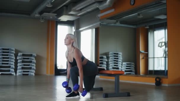 Mulher esportiva e loira se exercitando no clube de fitness fazendo uma perna sentar-se enquanto segurando halteres de cor azul. Vista lateral — Vídeo de Stock