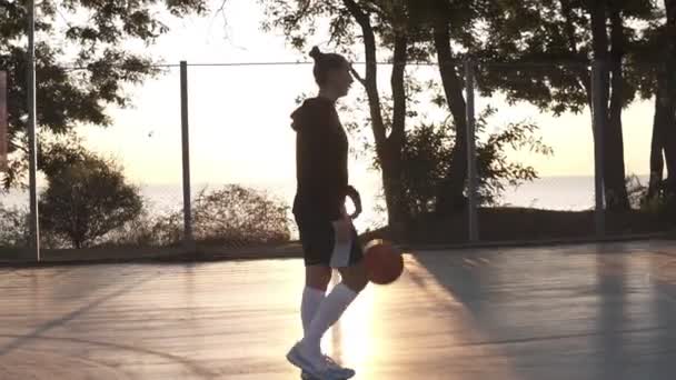 Jong meisje basketballer wandelen door basketbalveld buiten in de ochtend tijdens het stuiteren van de bal. Zijaanzicht — Stockvideo
