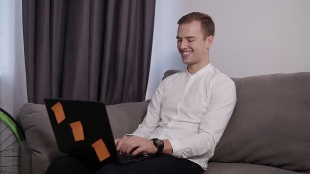 Расслабленный, улыбающийся молодой человек, сидящий дома на диване и работающий над ноутбуком. Счастливого настроения, фрилансер. Номер с современным интерьером в серых и белых тонах — стоковое видео