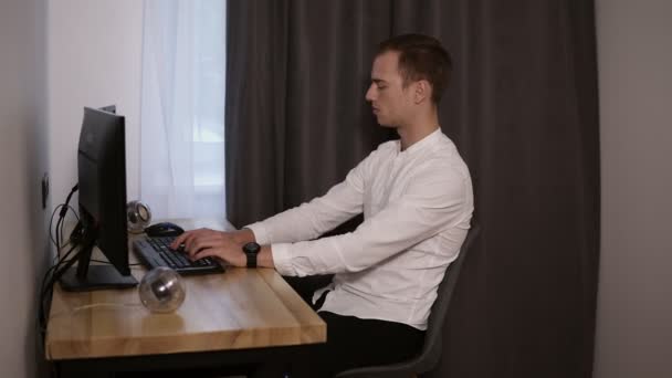 Ung man frilansare arbetar hemma, sitter vid skrivbord, att skriva på tangentbordet, använda en mus. Mannen i vit skjorta avslappnad och leende sitter framför skärmen copmuter. Sidovy — Stockvideo