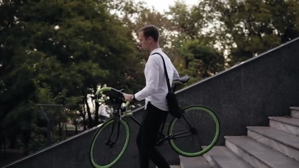 Bonito jovem homem de camisa branca e calças pretas está carregando um gren trekking e bicicleta de cor preta enquanto desce ao ar livre — Vídeo de Stock
