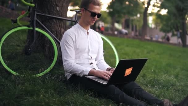 Joven empresario exitoso en camisa blanca y gafas negras. Hombre sonriente sentarse en el suelo de hierba, trabajar en el ordenador portátil en el parque de la ciudad en el césped verde al aire libre en la naturaleza se apoya en su bicicleta limpia el árbol — Vídeo de stock