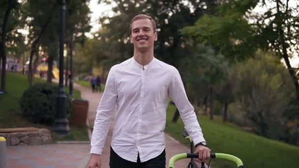 白人微笑着穿着白色衬衣的年轻人在城里的街上骑着自行车走路。在公园散步时滚动他的徒步自行车。前视图 — 图库视频影像