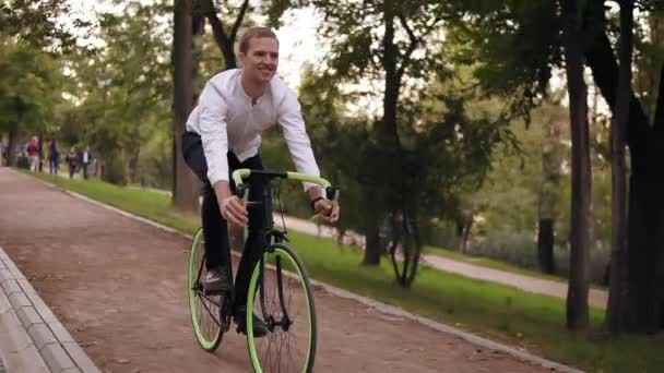 Счастливый, улыбающийся молодой человек в белой рубашке катается на велосипеде по тропинке в зеленом городском парке. Езда на велосипеде во время прогулки по парку — стоковое видео