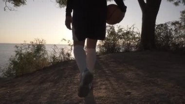 Bir basketbol oyuncu kız ile topu elinde, etrafındaki ağaçlarla ile eğim için geliyor geliyor. Denizin üzerinde parlayan güneş bakar. Deniz manzaralı. Arka görünüm