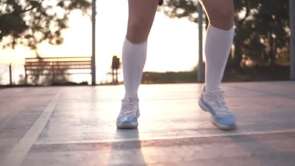 Dekat dengan kaki pemain basket wanita di kaus kaki golf putih yang melakukan olahraga dribbling dengan sangat cepat, berjalan mundur, melatih di luar ruangan di lapangan lokal. Matahari bersinar di latar belakang — Stok Video