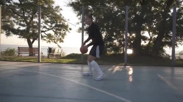 Side footage of a young girl basketball player training and exercising outdoors on the local court. Dribbling with the ball, bouncing and make a shot — Stock Video