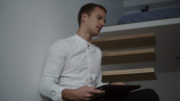 Вид збоку молодого професійного фрілансера в білій сорочці з використанням портативного комп'ютера, сидячи на сучасних сходах. Знімок під низьким кутом — стокове відео