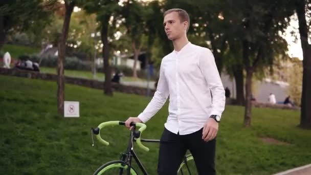 Jovem caucasiano de camisa branca andando de bicicleta na rua da cidade. Rolando sua bicicleta de trekking enquanto caminhava pelo parque pensativo — Vídeo de Stock