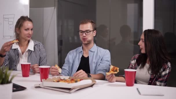 Junge kaukasische Mitarbeiter, die in der Mittagspause im Büro gemeinsam Pizza essen. Lachen, reden, Spaß haben. Essen, Business Lunch und People Konzept - glückliches Kreativteam isst Pizza im Büro — Stockvideo