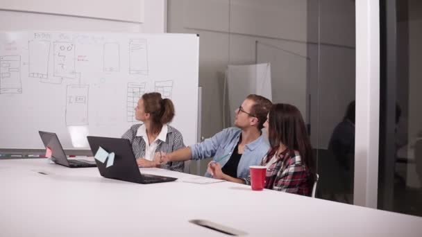 Homem jovem em óculos e duas mulheres em casual sentado na mesa de escritório no local de trabalho criativo com laptop nele. Homem no meio apontando com lápis no quadro branco, explicando sua visão ou — Vídeo de Stock