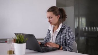 Kız at kuyruğu ile modern loft studio dizüstü bilgisayar kullanın. İşlem çalışma araştırma, yazarak. Genç iş kadını ile yaratıcı başlangıç çalışma. Küçük yeşil çiçek ve arkasında kum saati belgili tanımlık laptop. Yavaş
