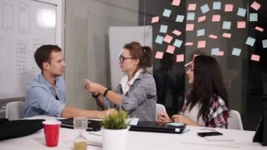 Gençler olumlu birlikte yapışkan renkli kağıtlar ile cam duvarın arkasında büyük beyaz ofis masada oturan beyin fırtınası sırasında çalışırken gülüyor. Neşeli speople rahat öğrenme