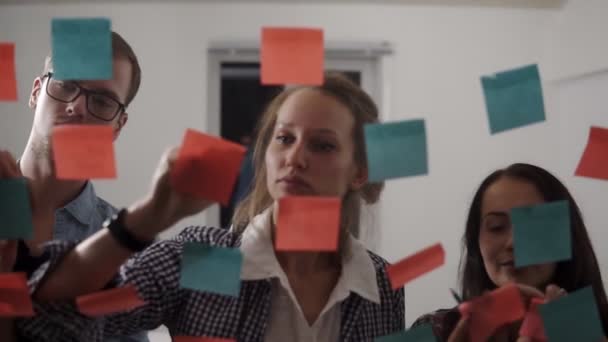 Das Team besteht aus drei jungen Leuten - jungen Geschäftsleuten, die beim Brainstorming zusammenarbeiten und hinter Glaswänden mit klebrigen bunten Papieren stehen. Frontansicht — Stockvideo