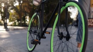 Pedallar itmek bir genç kız kot pantolon ve spor ayakkabı yeşil tekerlekli, trekking bisiklet sürme başlangıç görüntüleri kapatın. Kent manzaralı, Mercek Parlaması, yüzü yok. Odaklanmamış arka plan