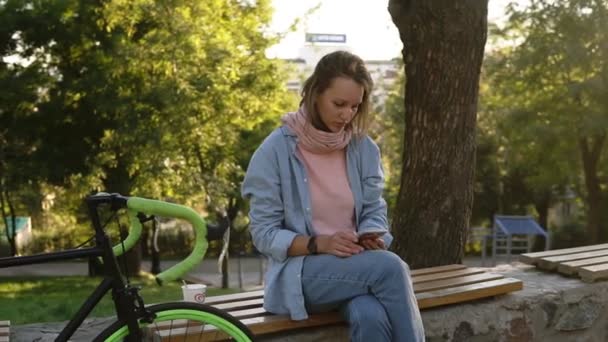 Chica pensativa sentada en el banco en el parque de la ciudad con su bicicleta de trekking a su lado. Mirando su móvil, vistiendo ropa casual brillante. Árboles y sol en el fondo — Vídeos de Stock