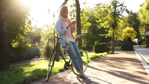 Ελκυστική κοπέλα κλίνει σχετικά με το ποδήλατο στο πάρκο της πόλης. Κοιτάζοντας της κινητής τηλεφωνίας, φορούν φωτεινά ροζ και μπλε χρωματιστά casual ρούχα. Δέντρα και τον ήλιο στο φόντο. Επισκόπηση — Αρχείο Βίντεο